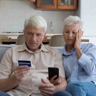 A senior couple with alarmed looks on their faces look at a cell phone and credit card.