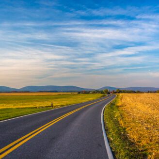 A two lane road lined with fields going off towards mountains