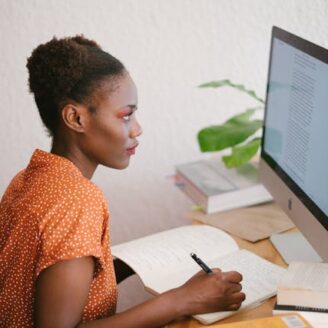 woman looks at a computer screen while writing in a notebook at her desk
